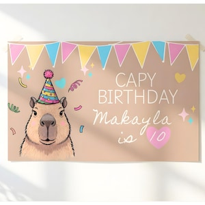 Op de afbeelding: Een beige verjaardagsbanner met een cartoon capibara met een feesthoed. De banner leest "CAPY BIRTHDAY Makayla is 10" met kleurrijke confetti en slingers. Een roze hart staat naast het nummer 10.