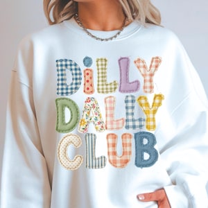 Puede incluir: Una sudadera blanca de manga larga con el texto DILLY DALLY CLUB en letras grandes y multicolores con aspecto de tela. Cada letra muestra un patrón diferente, incluyendo cuadros vichy azules, lunares rojos, cuadros verdes, flores y rayas.