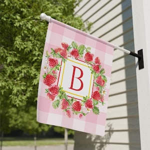 May include: A pink and white gingham garden flag with a wreath of red strawberries and green leaves surrounding a yellow square with the letter "B" in red.