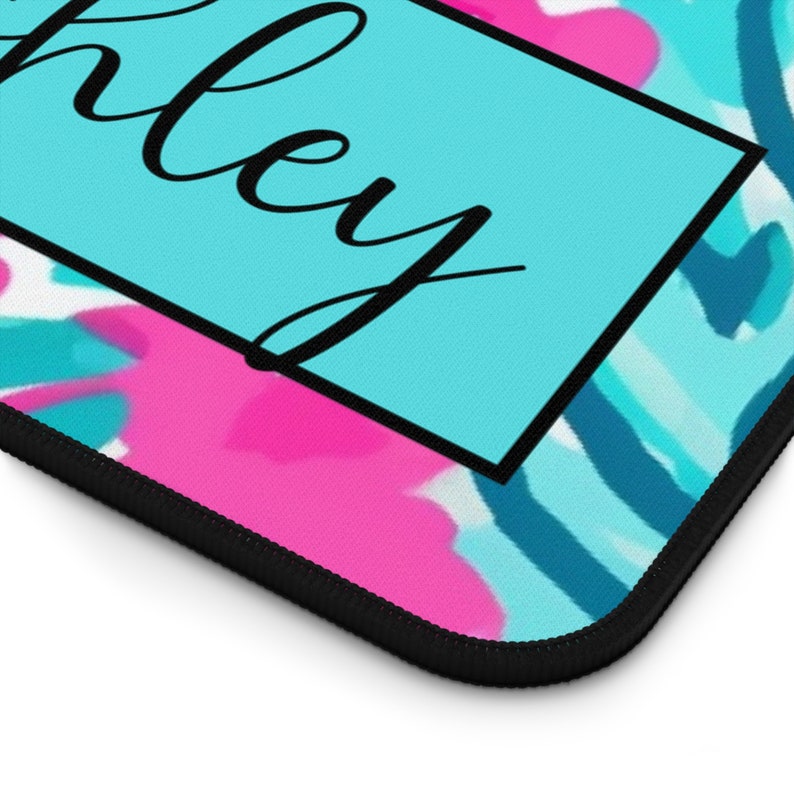 May include: A blue and pink abstract patterned mousepad with a black border and the word "Ashley" written in black cursive inside a white box.