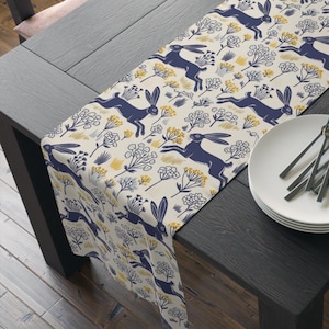 May include: A cream-colored table runner featuring a repeating pattern of navy blue hares and floral elements in yellow and blue. The runner is draped over a dark wooden table, with a stack of white plates and silverware visible.