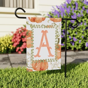 Peut inclure: Un drapeau de jardin avec la lettre "A" orange au centre, entouré de citrouilles à l'aquarelle et de vignes vertes. Le drapeau est sur un support en métal noir, sur fond d'herbe verte et de fleurs colorées. La hauteur du drapeau est d'environ 30-40 cm.
