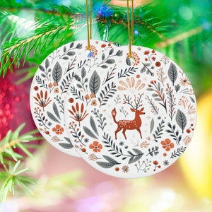 May include: White ceramic Christmas ornament with a red deer and floral pattern. The ornament is round and has a gold string for hanging.