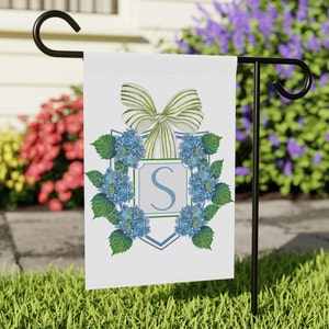 May include: A white garden flag with a blue and green hydrangea wreath and a white bow with green stripes. The wreath surrounds a blue shield with the letter "S" in the center.