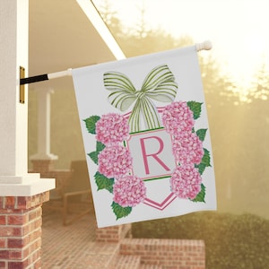 May include: White garden flag featuring a pink floral wreath with the letter "R" in the center. A green and white striped bow is at the top. The flag is hanging outside on a sunny day.