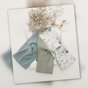 Könnte beinhalten: Drei Stoff-Haarbänder in Pastellfarben. Eines ist gerippt blau, ein anderes strukturiert grün und das dritte weiß mit einem Eukalyptusblatt-Druck. Die Haarbänder sind geknotet und auf einer weißen Oberfläche angeordnet.
