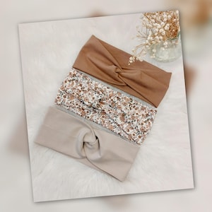 Könnte beinhalten: Drei Stirnbänder in verschiedenen Farben und Mustern. Das obere Stirnband ist einfarbig braun, das mittlere hat ein Blumenmuster und das untere ist einfarbig beige. Alle drei haben einen gedrehten Knoten.