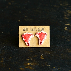 Peut inclure: Une paire de boucles d'oreilles en forme de steak, avec le texte "Well that's bizarre" sur une carte marron.
