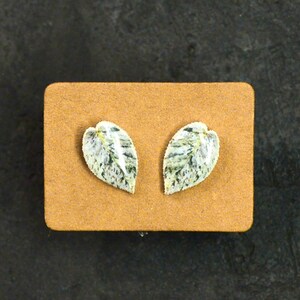May include: A pair of leaf-shaped earrings. Each earring features a white base with green and gray veining, resembling a leaf. The earrings are displayed on a small, rectangular, tan-colored card against a dark background.