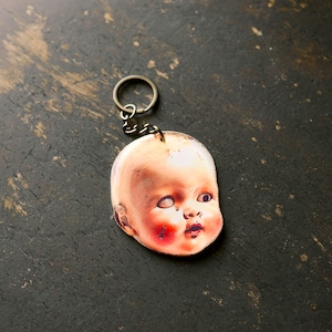 May include: A vintage-style doll head keychain with a painted face and a distressed white finish. The doll has blue eyes, rosy cheeks, and a slightly open mouth. The keychain is attached to a silver metal ring.
