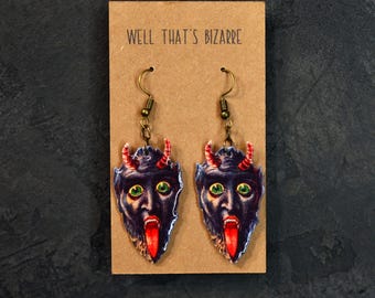 May include: A pair of dangle earrings featuring a cartoon devil face with green eyes, a red tongue, and red horns. The earrings are made of wood and have a gold-tone hook.