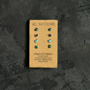 May include: A card with the text "WELL THAT'S BIZARRE" and "STRING OF THINGS:" with a list of items: "String of Hearts", "Varlegated String of Hearts", "Stiver Glory String of Hearts", "String of Turtles". The card has six pairs of small heart-shaped earrings with green, pink, and blue designs.