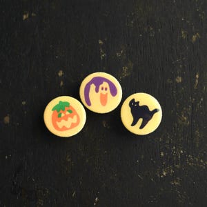 Puede incluir: Tres botones amarillos con temática de Halloween con diferentes diseños. Un botón tiene un fantasma morado, otro tiene un gato negro y otro tiene una calabaza naranja.