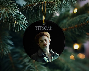 May include: A round, black Christmas ornament with a gold string for hanging. The ornament features a portrait of a person with the text "TITSOAK" above the image. The background is a dark color.