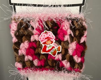 Könnte beinhalten: Handgemachte Kaffeebecher-Manschette mit einem Häkeldesign in Rosa- und Brauntönen. Die Manschette ist mit einem Erdbeerkuchen-Aufkleber verziert und mit flauschigem rosa Garn verziert. Entworfen für eine Standard-Kaffeetasse.