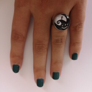 May include: A silver ring with a black and white glass cameo featuring a silhouette of Jack Skellington and Sally from the Nightmare Before Christmas movie. The ring is on a hand with dark green nail polish.