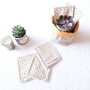 May include: Three white fabric squares with a black and gold geometric pattern, a small potted succulent plant, and a white candle in a glass jar with a black and gold geometric pattern.