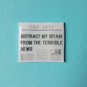 May include: A white rectangular sticker with black text that reads "THE 1975 DISTRACT MY BRAIN FROM THE TERRIBLE NEWS".