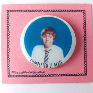 May include: A round blue and white button with a photo of a person wearing a white shirt and a black and white striped tie. The text "COMPLETED IT MATE" is printed on the button.