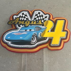 May include: A blue and yellow cake topper featuring a cartoon race car with the number 43 on the side. The car is surrounded by checkered flags and the name "Angus" is written in gold letters. The number 4 is also featured in gold letters.