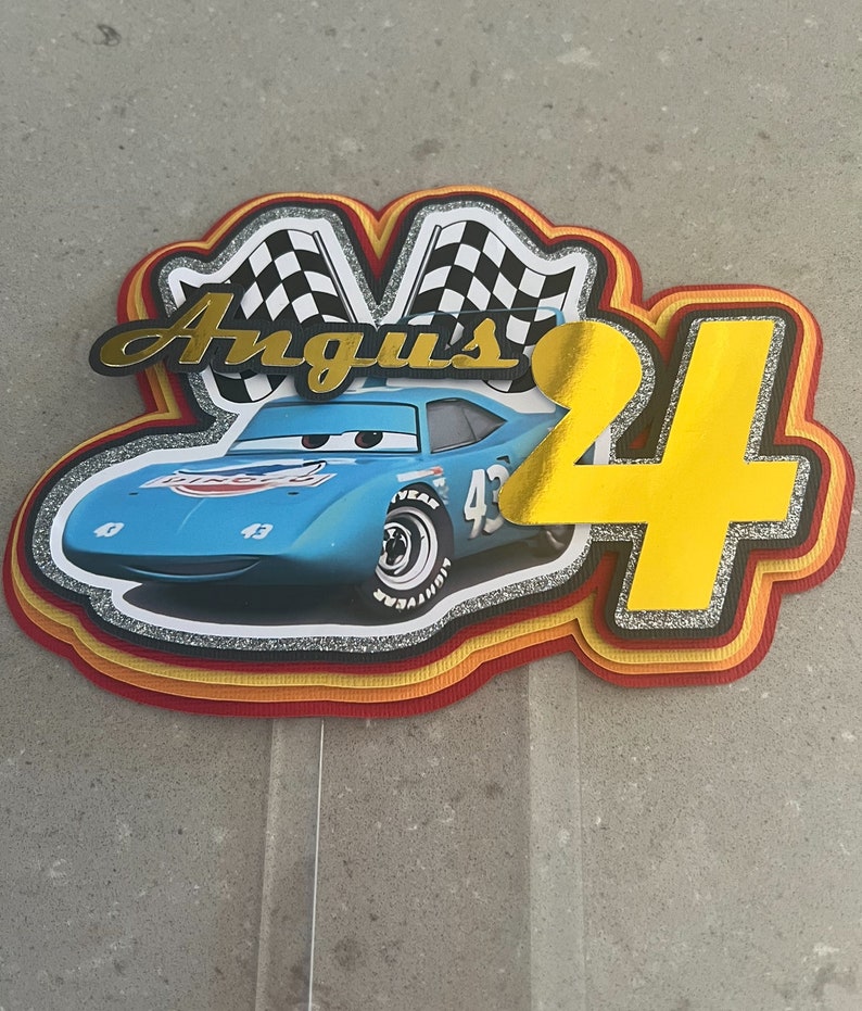 May include: A blue and yellow cake topper featuring a cartoon race car with the number 43 on the side. The car is surrounded by checkered flags and the name "Angus" is written in gold letters. The number 4 is also featured in gold letters.