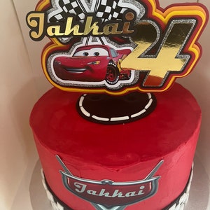 May include: Red and gold Cars themed birthday cake with a checkered flag topper, a red race car, and the name "Jahkai" written in gold. The cake is decorated with a checkered flag pattern and a red Cars logo.
