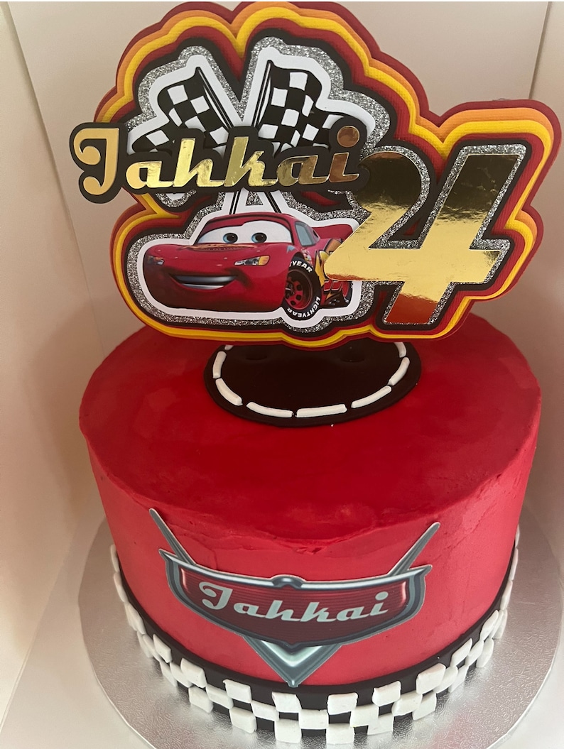 May include: Red and gold Cars themed birthday cake with a checkered flag topper, a red race car, and the name "Jahkai" written in gold. The cake is decorated with a checkered flag pattern and a red Cars logo.