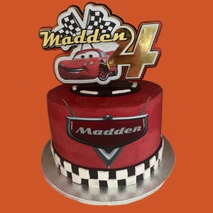 May include: A red and black Cars themed birthday cake with a red race car on top. The cake is decorated with a checkered flag design and the name "Madden" is written on the cake.