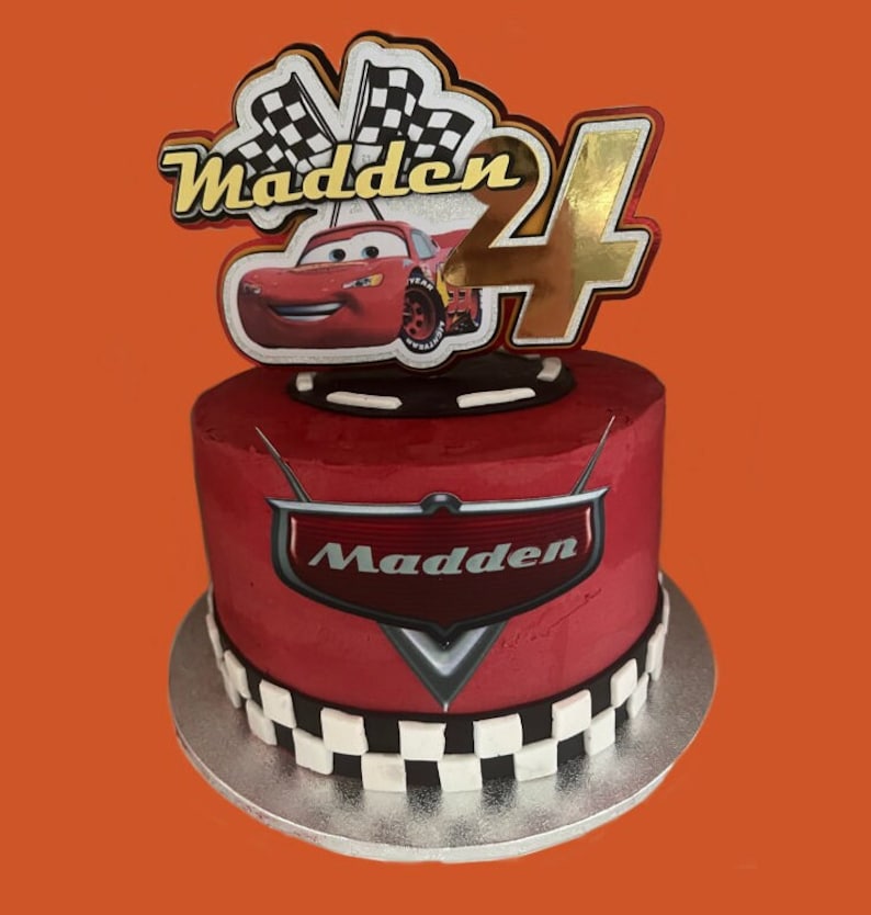 May include: A red and black Cars themed birthday cake with a red race car on top. The cake is decorated with a checkered flag design and the name "Madden" is written on the cake.