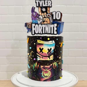 May include: A Fortnite-themed cake featuring a Marshmello character design. The cake is decorated with colourful sprinkles and a topper with the name "Tyler" and the number "10". The cake is on a white cake stand.