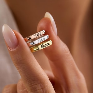 May include: Three personalised rings in rose gold, silver, and gold are held in a hand. Each ring has a name or symbol engraved on it: "Love", "Lella B", and "Grace". The rings are simple bands, perfect for stacking.