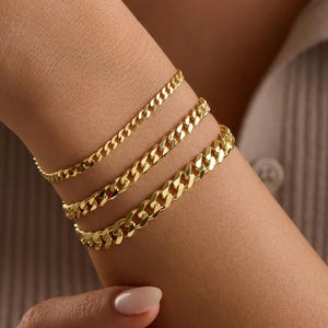 May include: Three gold-colored chain bracelets of varying widths are stacked on a wrist. The bracelets have a curb chain design, with a polished finish. The skin tone is light, and the background is a neutral color.