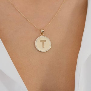 Op de afbeelding: Een gouden ketting met een ronde hanger met de letter "T" in een cirkel van kleine diamanten.