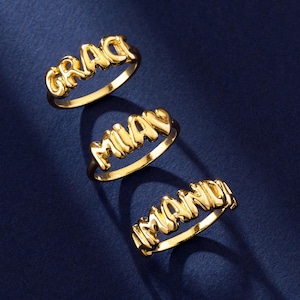 May include: Three gold-colored name rings spelling out "GRACE", "MIA", and "MANDY". The rings are displayed on a dark blue surface, with the names in a bubbly font.