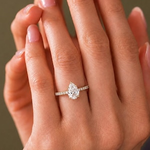 May include: A gold ring featuring a pear-shaped center stone and a band accented with small round stones. The ring is worn on a finger, showcasing the clear, faceted gemstone set in a prong setting with a thin, polished gold metal band.