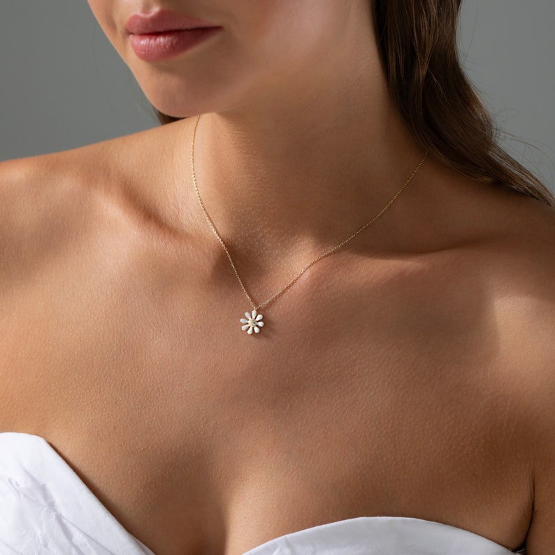 May include: A delicate gold chain necklace with a small white daisy pendant. The pendant is made of metal and has a slightly textured surface.