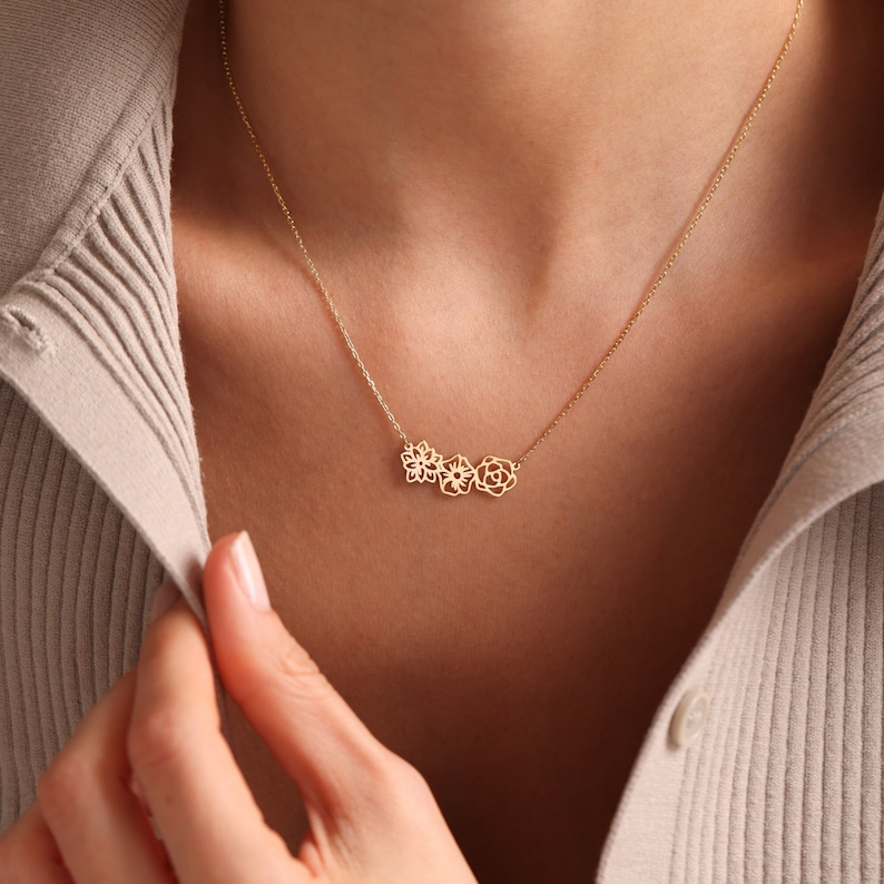 K&ouml;nnte beinhalten: Eine goldene Halskette mit einer zarten Kette und drei kleinen, ausgeschnittenen Blumenanh&auml;ngern.