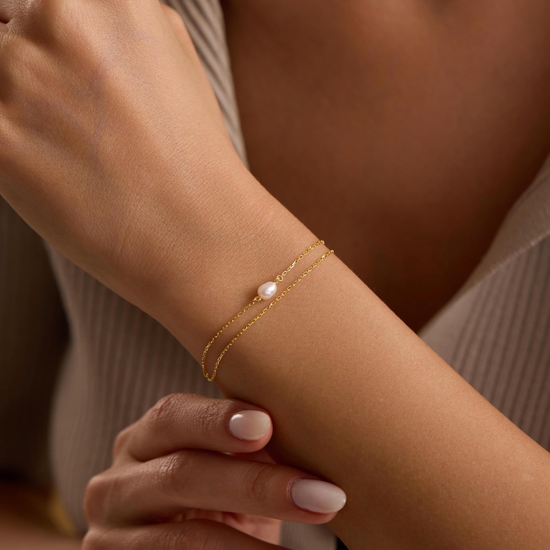 May include: A delicate gold bracelet featuring a double chain design. A single, lustrous white pearl is centered on the bracelet. The jewelry is displayed on a person's wrist, showcasing the elegant simplicity of the piece.