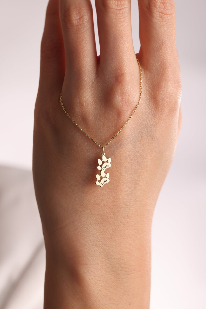 May include: Gold necklace with a paw print charm. The charm has the names "Daisy" and "Sadie" engraved on it.