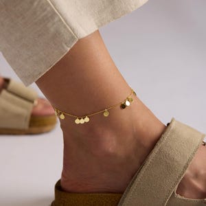 May include: A gold-colored anklet with small, round disc charms. The delicate chain is draped around an ankle, with a neutral-toned sandal visible below. The anklet adds a touch of elegance to the casual look.