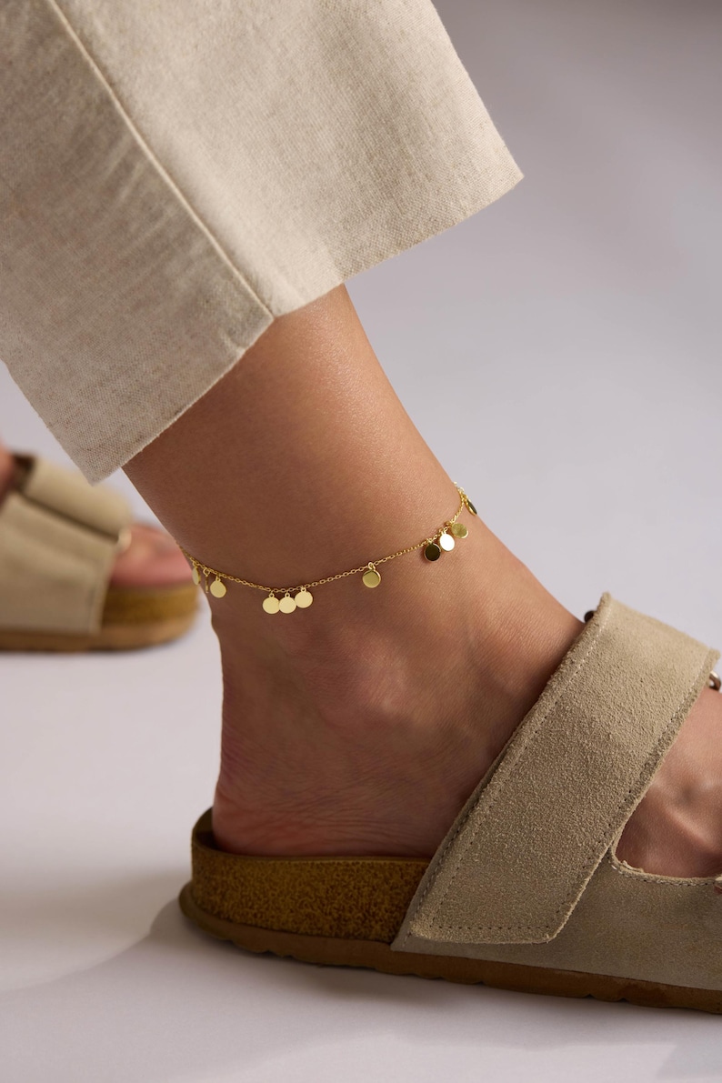 May include: A gold-colored anklet with small, round disc charms. The delicate chain is draped around an ankle, with a neutral-toned sandal visible below. The anklet adds a touch of elegance to the casual look.