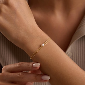 May include: A delicate gold bracelet featuring a single, round pearl. The bracelet is a simple design with a fine chain, displayed on a person's wrist. The background is a neutral color, highlighting the jewelry.