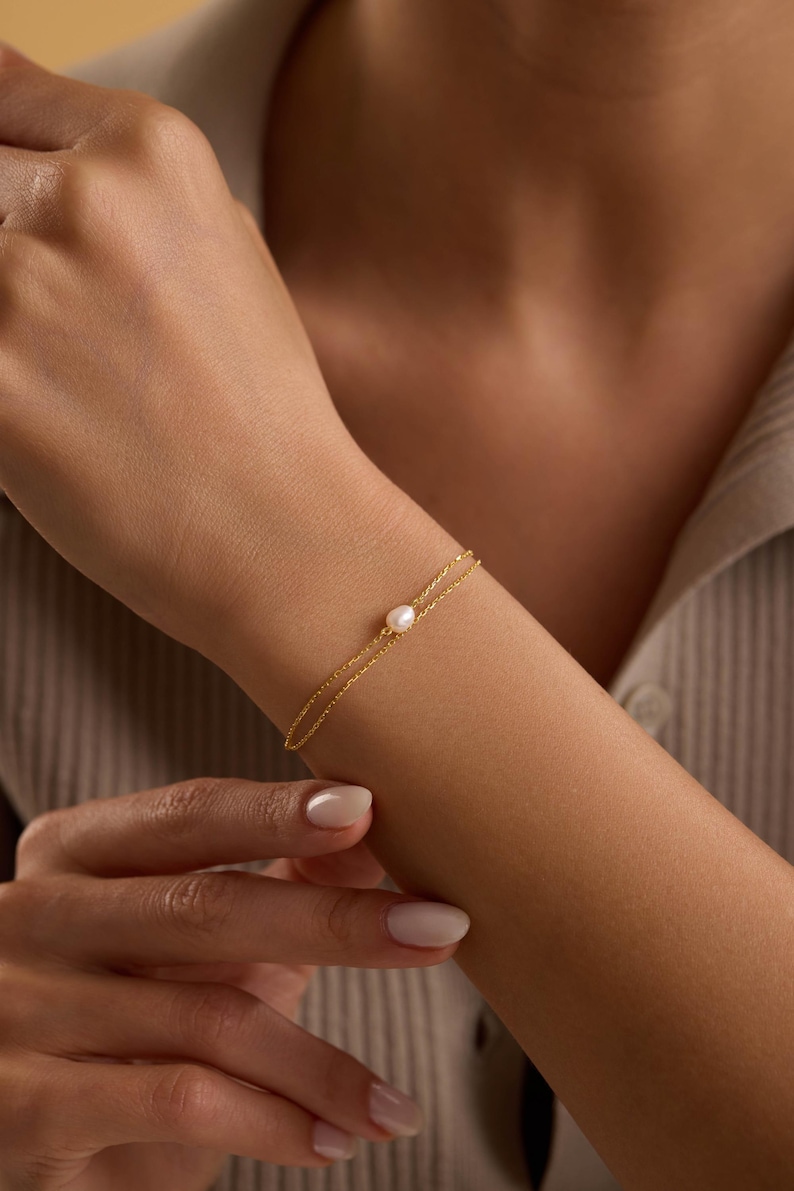 May include: A delicate gold bracelet featuring a single, round pearl. The bracelet is a simple design with a fine chain, displayed on a person's wrist. The background is a neutral color, highlighting the jewelry.