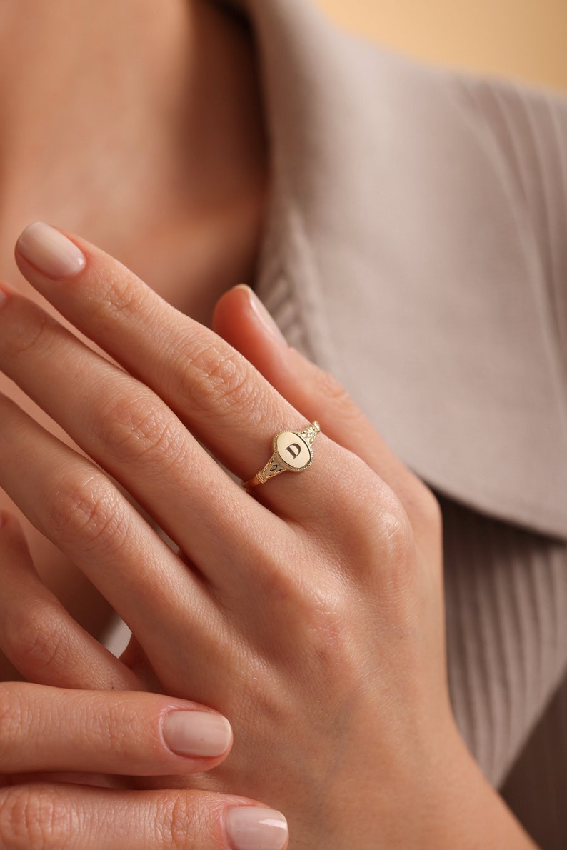 May include: A gold signet ring with an oval-shaped top featuring the letter "D" engraved in the center.