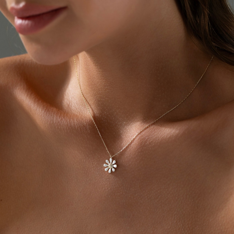 May include: A delicate gold chain necklace with a small white daisy pendant. The daisy has a yellow center and is accented with tiny sparkling stones.