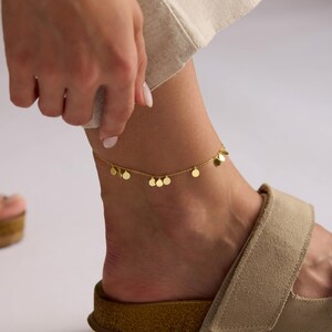 May include: A gold-colored anklet with small, dangling circular charms. The delicate chain is worn on an ankle, paired with a tan sandal. The image is set against a neutral background.