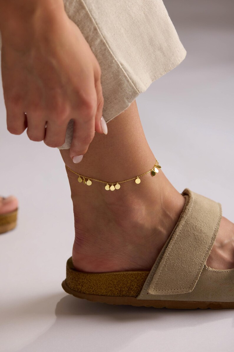 May include: A gold-colored anklet with small, dangling circular charms. The delicate chain is worn on an ankle, paired with a tan sandal. The image is set against a neutral background.