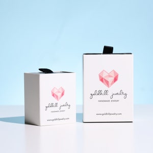 May include: Two white boxes with black ribbon handles. Both boxes have a pink geometric heart design and the text "goldhill jewelry handmade jewelry www.goldhilljewelry.com"