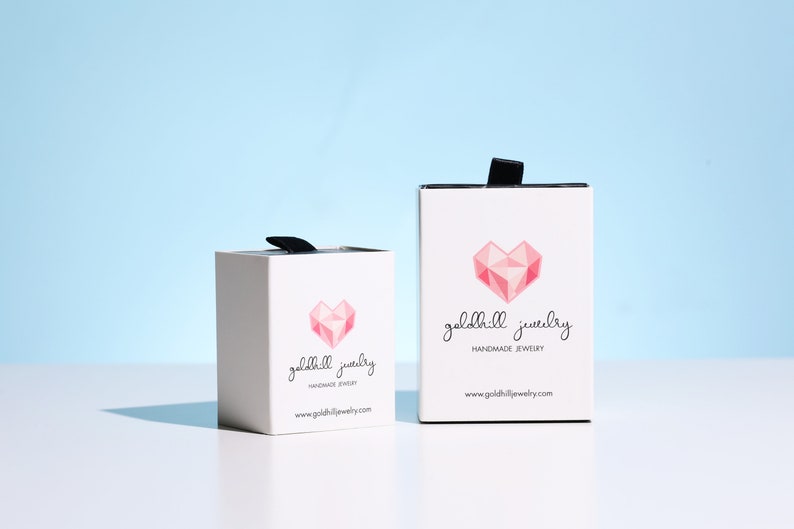 May include: Two white boxes with black ribbon handles. Both boxes have a pink geometric heart design and the text "goldhill jewelry handmade jewelry www.goldhilljewelry.com"