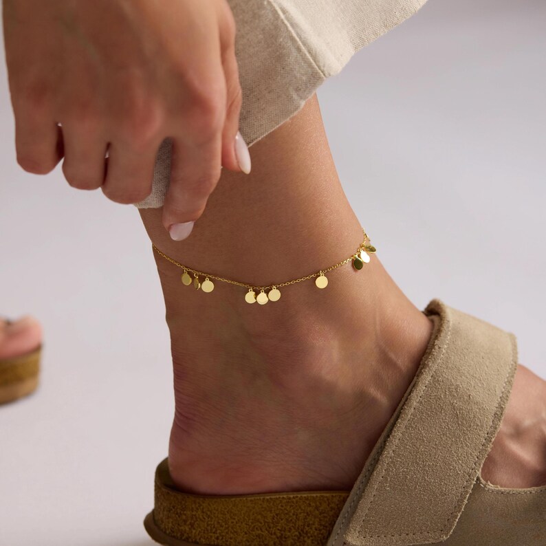 May include: A gold-colored anklet with small, dangling circular charms. The delicate chain is worn around the ankle, with the charms adding subtle movement. The anklet is shown on a person's ankle, paired with a sandal.